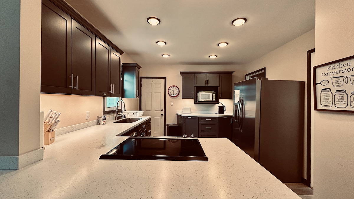 A modern kitchen is displayed with dark cabinetry and a central island featuring a smooth, white countertop. Essential appliances, including a microwave and refrigerator, are visible. Bright lighting enhances the open layout, while a wall displays kitchen conversion measurements for convenience.