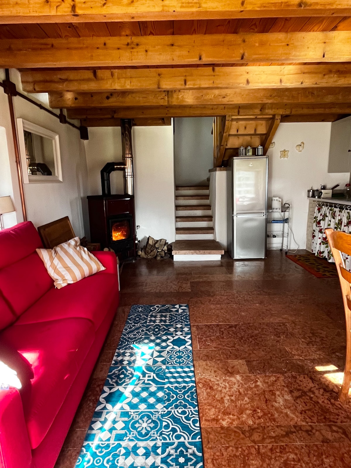 An inviting living area features a red sofa, a wooden beam ceiling, and a cozy wood-burning stove. A patterned rug extends across the floor, leading to a staircase that ascends to the upper level. A small kitchen area with a refrigerator is visible in the background.