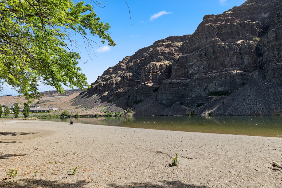 A serene riverside view is presented, featuring sandy banks that gently slope towards calm waters. Massive basalt cliffs rise in the background under a clear blue sky, with sparse greenery dotting the landscape. The tranquil scene conveys a sense of natural beauty and openness.