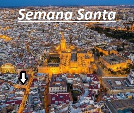 An aerial view captures the illuminated cityscape of Sevilla during Semana Santa. The prominent Cathedral and Giralda tower stand out, showcasing intricate architectural details. Surrounding buildings reflect warm lighting, creating a vibrant evening atmosphere. The narrow streets below are lined with traditional white houses, enhancing the local charm.