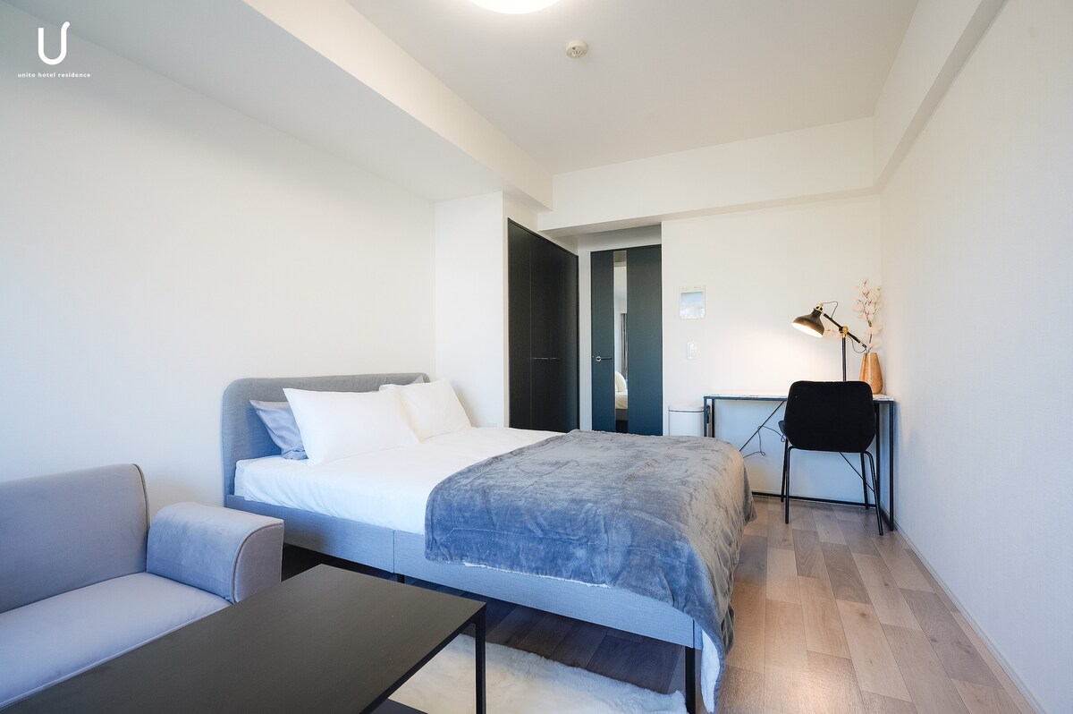 The spacious room features a comfortable bed adorned with soft linens. A desk with a chair is positioned nearby, while a sofa and low table create a cozy seating area. Light wood flooring complements the neutral-toned walls, contributing to a clean and modern aesthetic.