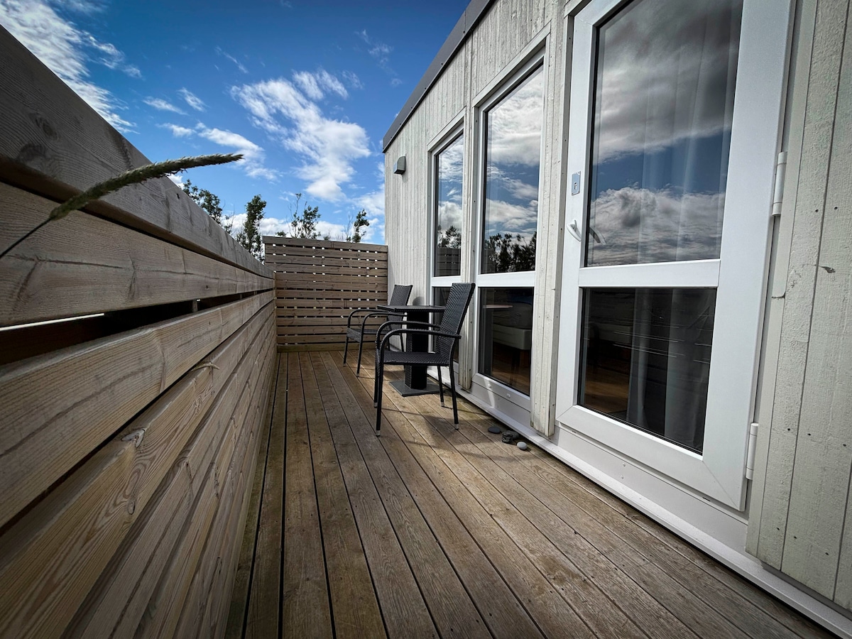 A wooden deck area presents two black chairs positioned beside large windows, allowing natural light to fill the space. The wooden walls provide a sense of privacy while surrounding greenery adds to the peaceful setting. Soft clouds drift across a blue sky above.