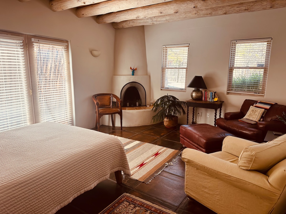 A spacious sleeping area features a queen-size bed covered with a light quilt. A cozy seating arrangement includes a leather armchair and a beige couch. Sunlight enters through two windowed walls, illuminating the room's warm tones, while a small fireplace is positioned at the center.