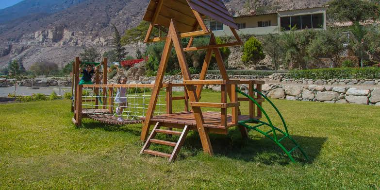 A wooden playground structure is situated on a grassy area, featuring swings and a climbing frame. The background showcases a scenic landscape with mountains and greenery. The space provides a safe environment for children to play and explore.