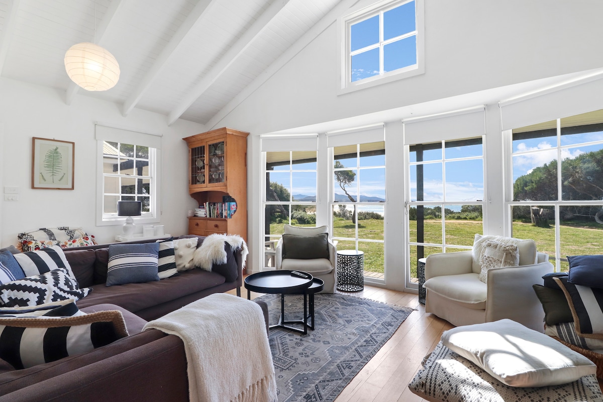 A spacious living area features large windows allowing natural light and views of the surrounding landscape. Cozy seating, including a dark sofa and armchairs, is arranged around a coffee table. A soft area rug adds warmth, complementing the light wood flooring.