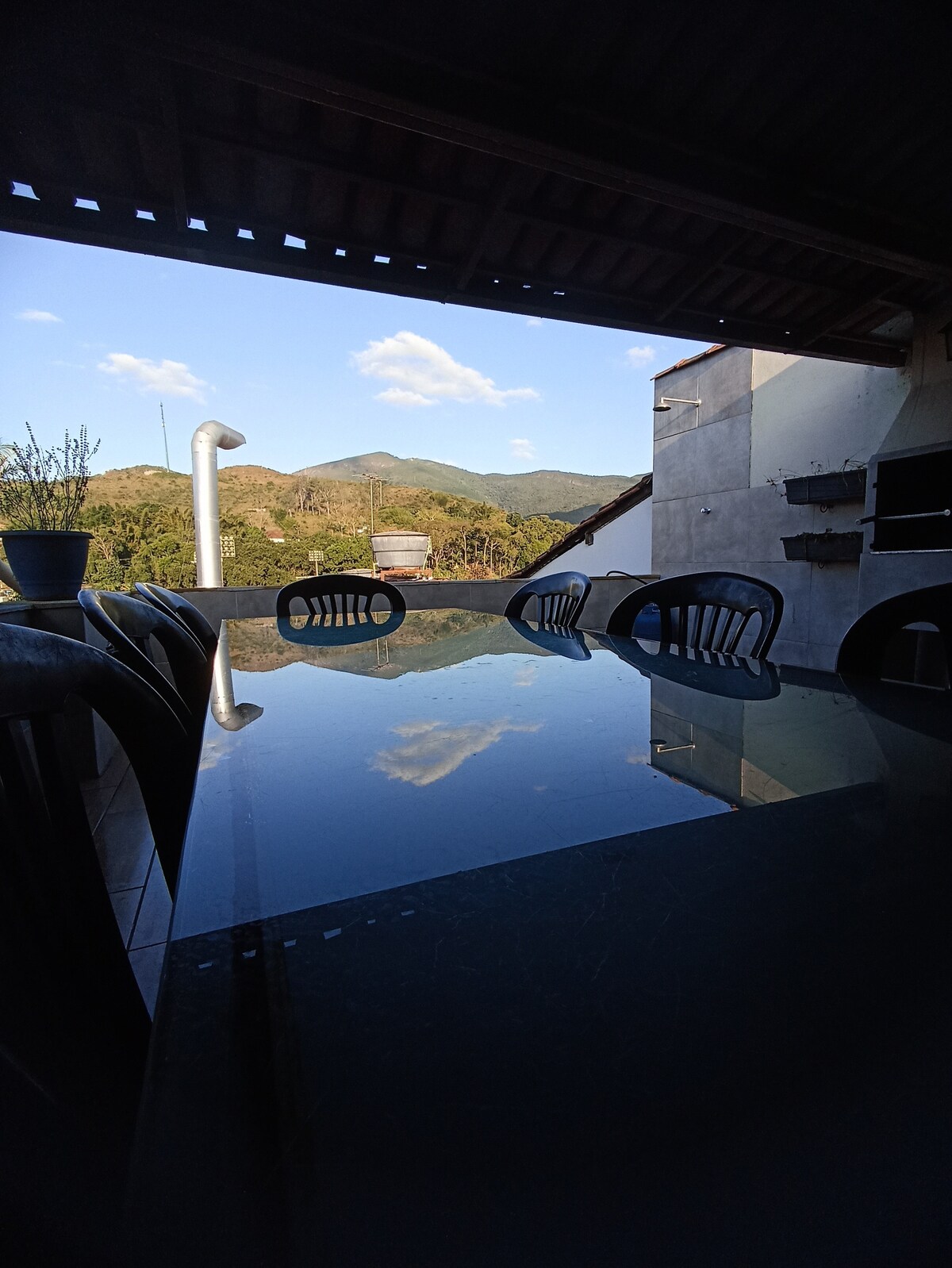 An outdoor dining area is depicted, featuring a glass table reflecting the surrounding landscape. Black chairs surround the table, and a distant mountain range can be seen in the background under a clear blue sky. A potted plant adds a touch of nature to the setting.