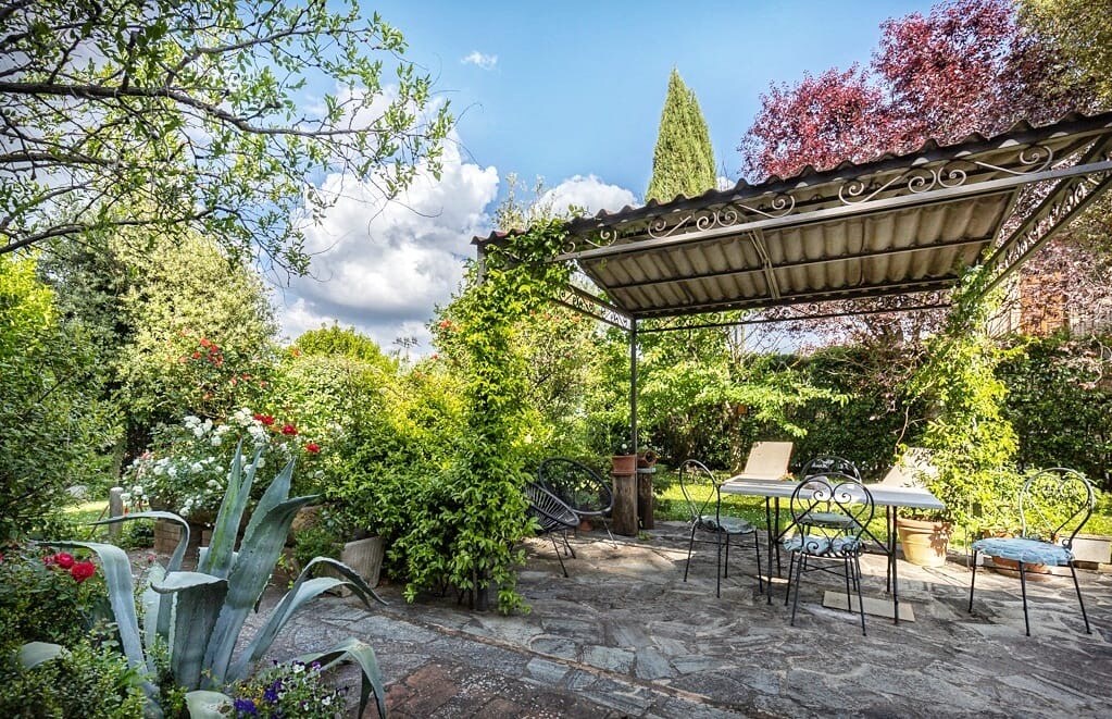 A serene outdoor seating area is surrounded by lush greenery and blooming flowers. A shaded pergola provides cover over a few metal chairs and a sun lounger, inviting relaxation. Soft textures of plants and trees create a peaceful environment for guests to enjoy.