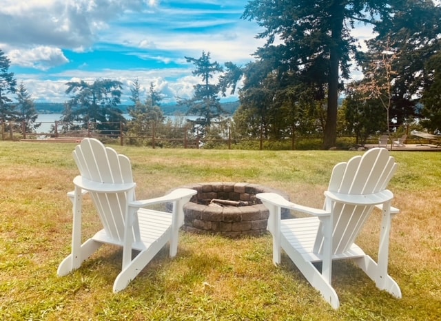 Two white Adirondack chairs are positioned around a circular stone fire pit on a grassy lawn. Beyond the seating area, a view of the water and surrounding trees adds to the outdoor ambiance.