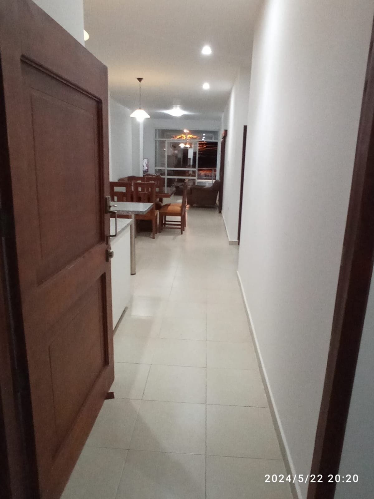 A well-lit hallway leads towards a dining area with a wooden table and chairs. Soft white walls and a tiled floor create a clean, bright ambiance. In the background, a living space is visible with additional seating and natural light entering through large windows.