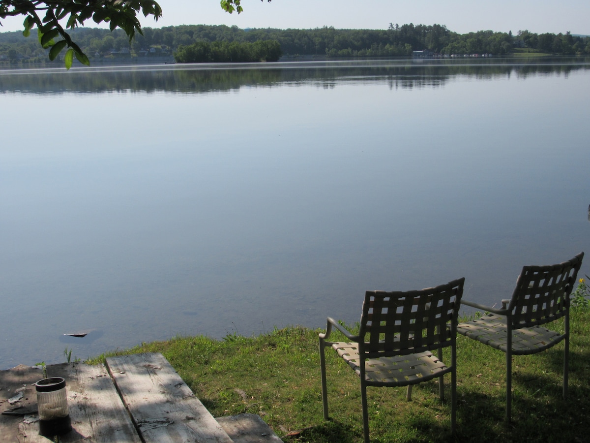 Two metal chairs are positioned on the grassy edge of a serene lake, offering a view of the calm water reflecting the surrounding lush greenery. A wooden table sits nearby, enhancing the outdoor setting.