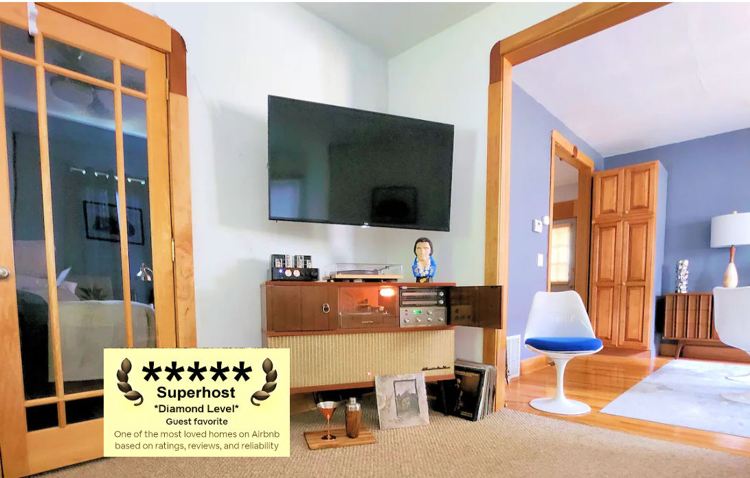 A modern living space is showcased, featuring a large wall-mounted TV above a mid-century modern stereo console. Light wood accents are present in the furniture and sliding glass door. Bright walls enhance the space, creating an inviting atmosphere for relaxation and entertainment.