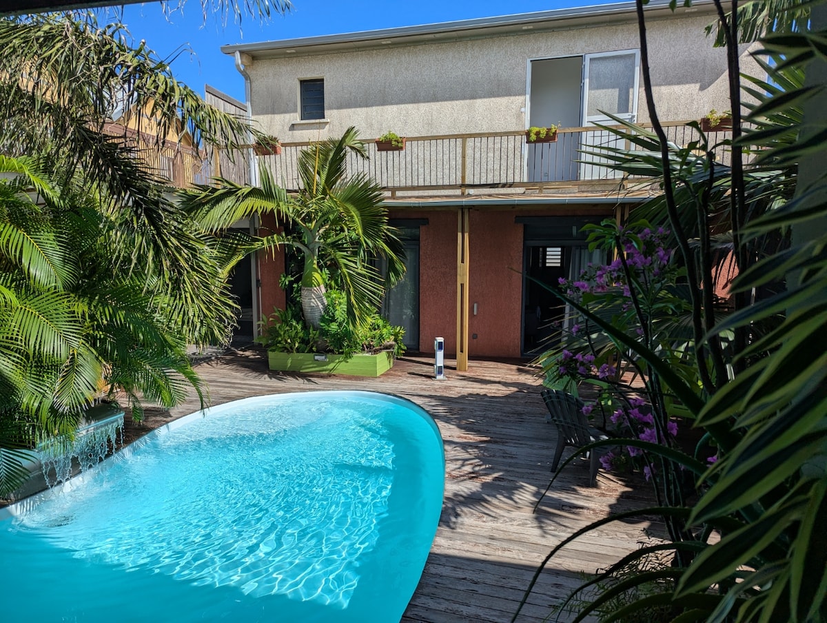 A serene pool is set within a lush garden space, surrounded by tropical plants and wooden decking. The patio is designed for relaxation, featuring one floor of the villa visible in the background. Bright sunlight enhances the inviting atmosphere of the area.