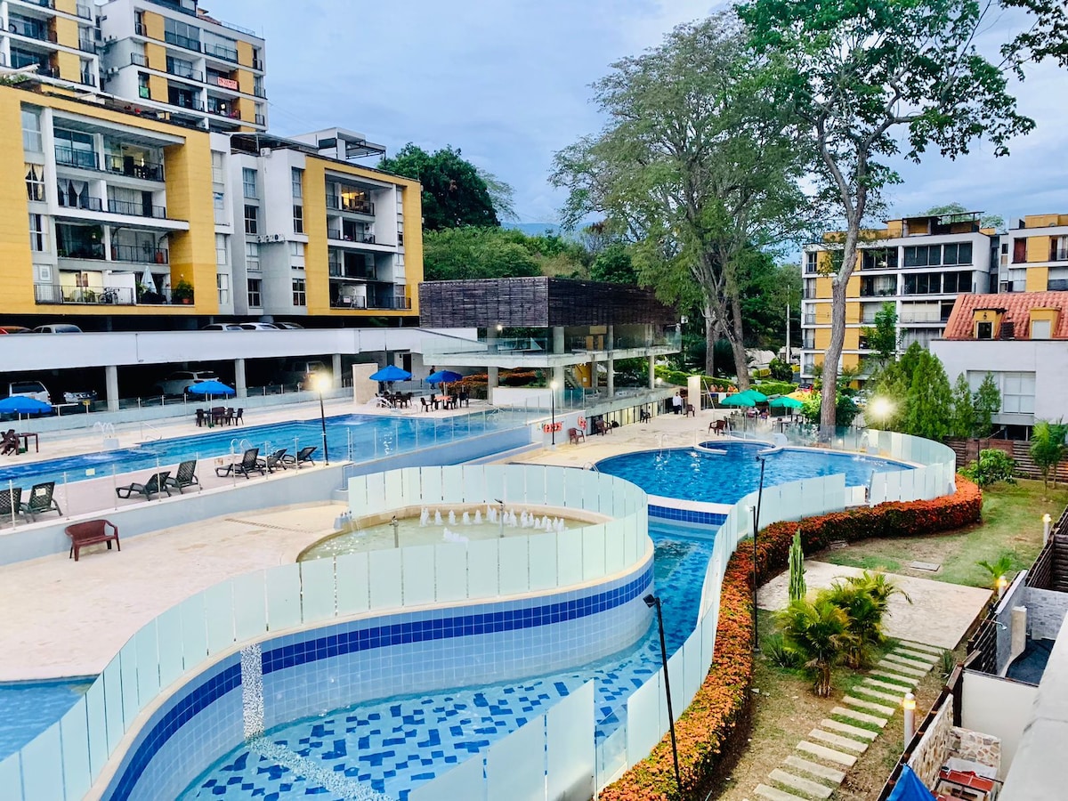 A spacious outdoor pool area is presented, featuring meandering pools separated by clear glass barriers. Sun loungers and umbrellas are positioned around the pools. Lush greenery surrounds the area, enhancing the tranquil environment created by the gentle water features.