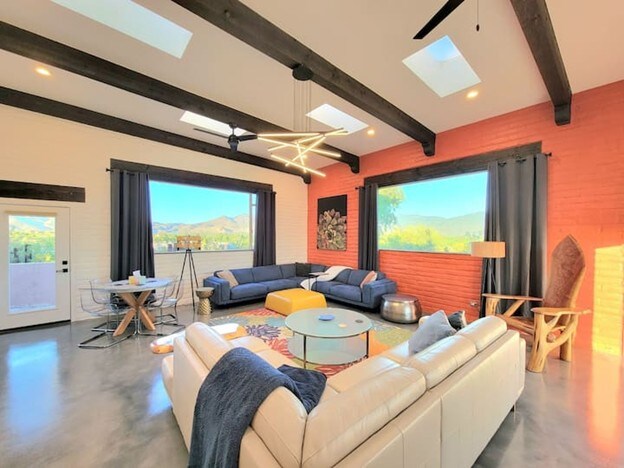 A spacious living area features a large sectional sofa arranged around a central coffee table. Natural light fills the room through multiple windows and skylights, highlighting modern artwork on the walls. Wooden beams add architectural interest, while a cozy corner includes a lamp and a decorative chair.