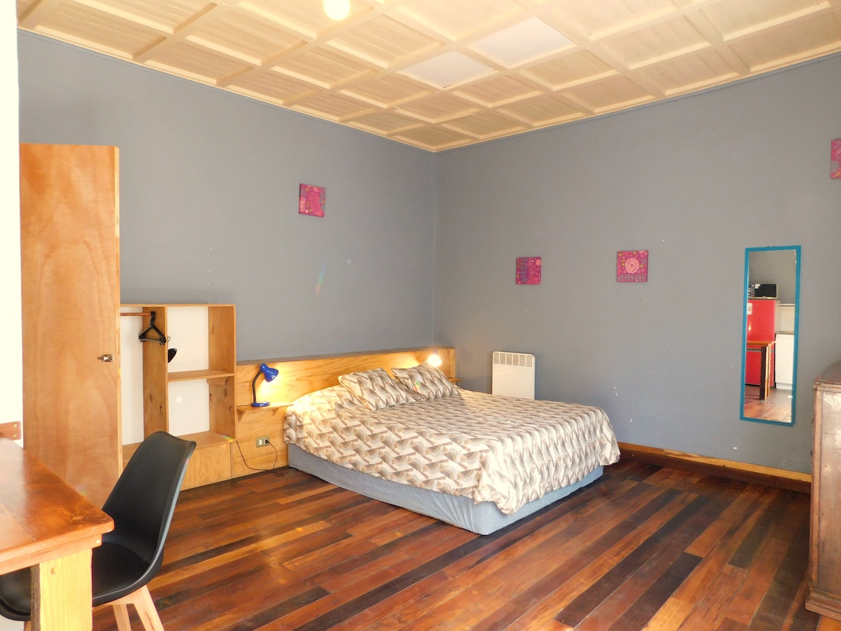 A spacious studio room features a large bed with a patterned coverlet against a blue wall. A small light desk is positioned nearby, and a mirror reflects the natural light in the room. Wooden flooring adds warmth to the space, while minimal decor includes framed art pieces.