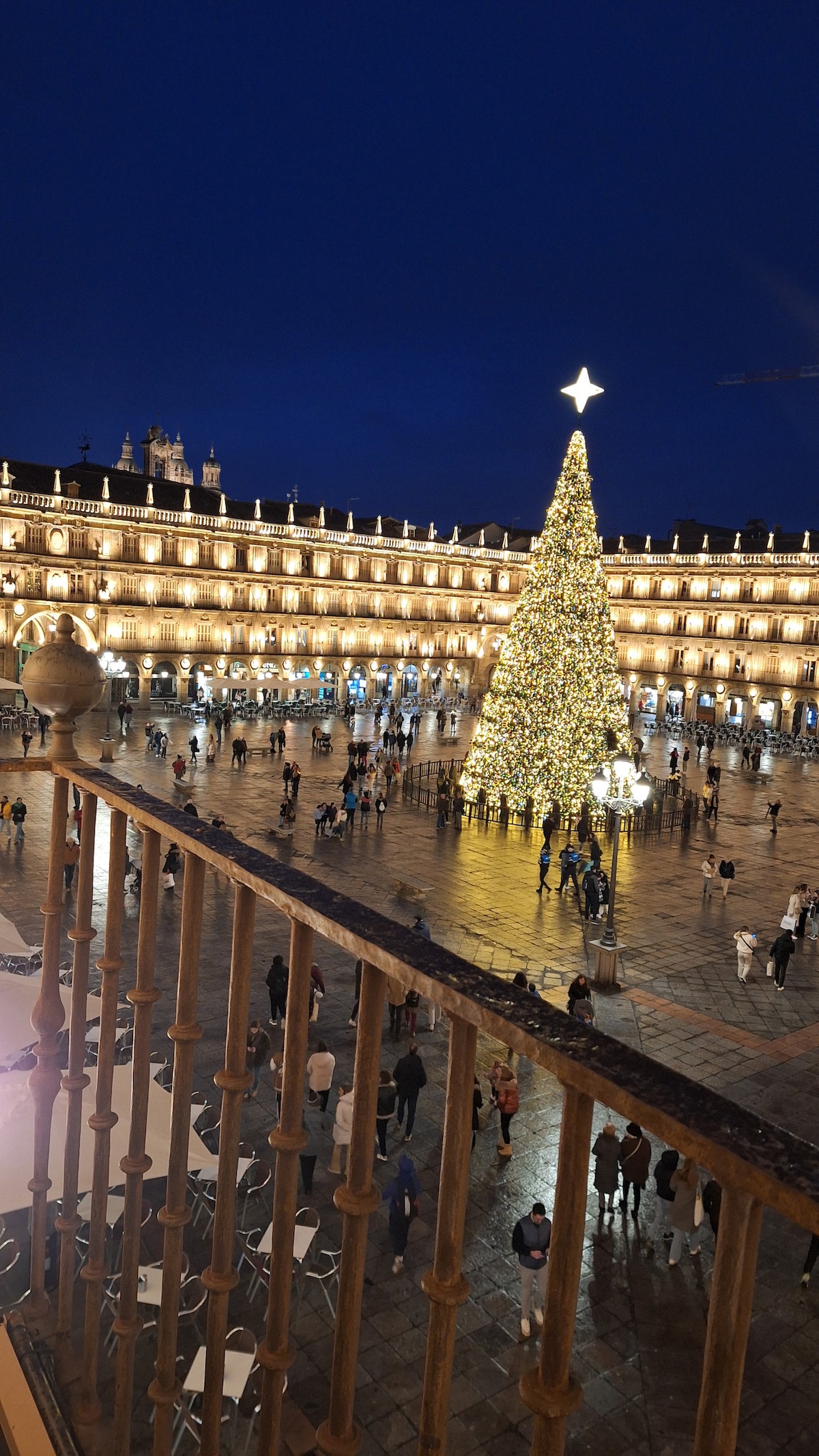 A nighttime view of Plaza Mayor is depicted, beautifully lit with festive decorations and a large Christmas tree at the center. The surrounding architecture is illuminated, highlighting its grandeur. People are seen walking and gathering throughout the plaza, creating a lively atmosphere.