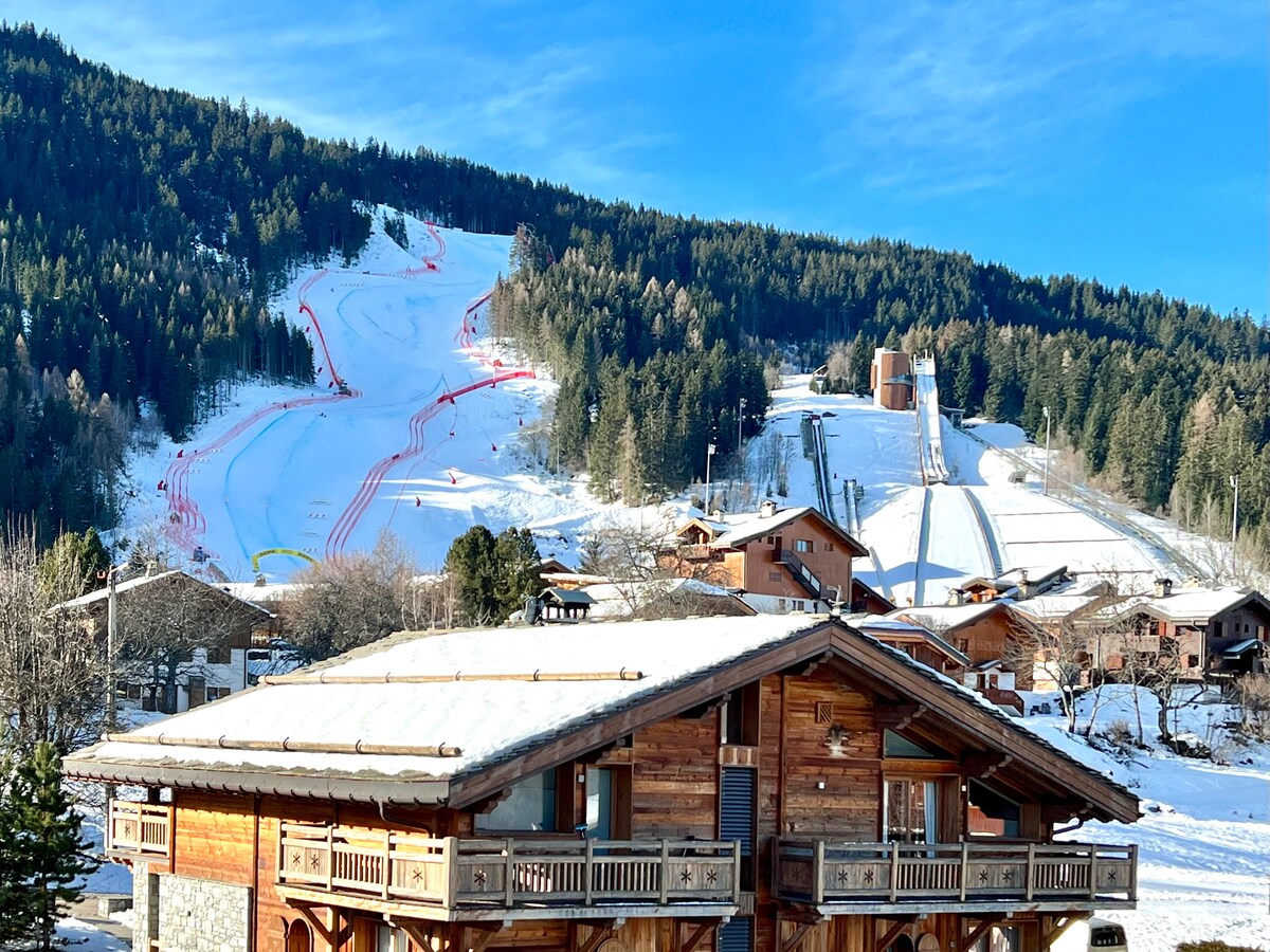 The image captures a charming chalet in the foreground, framed by snow-covered slopes in the background. Ski runs are visible, meandering down the mountain, while pine trees dot the landscape, contributing to a serene winter view. Blue skies provide a clear backdrop.