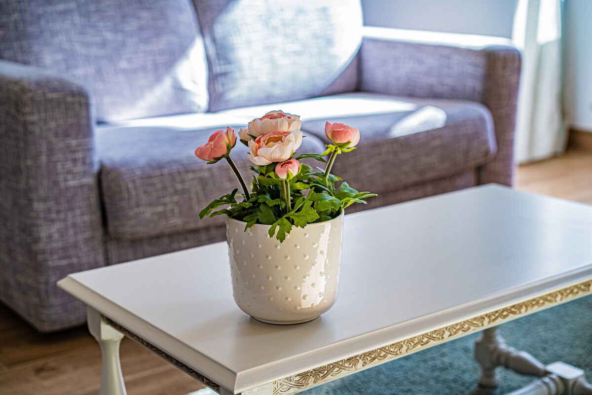 A potted plant with pink flowers is displayed on a white table. A gray sofa is visible in the background, providing a comfortable seating area. Soft natural light filters through a nearby window, creating a serene ambiance.
