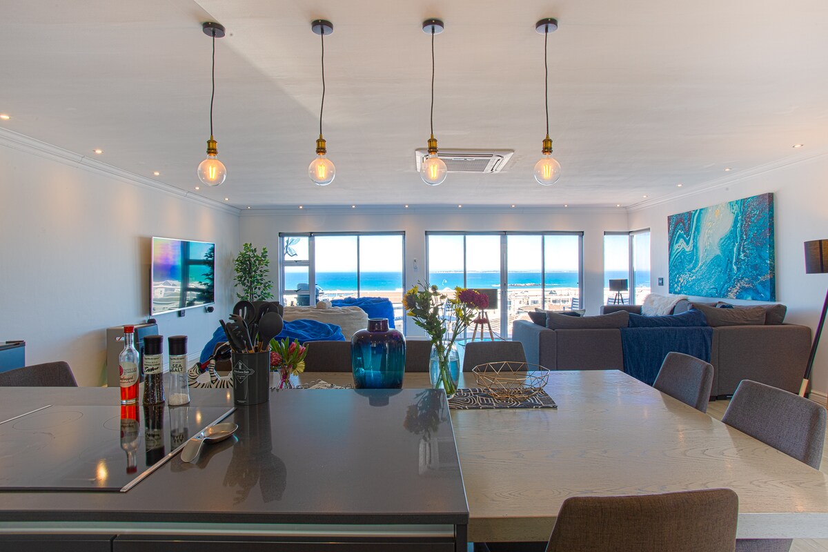 The open living space is highlighted by large windows offering expansive ocean views. A modern kitchen with a sleek island is visible in the foreground, complemented by a dining area. Soft furnishings and decorative art pieces provide a contemporary touch to the inviting atmosphere.