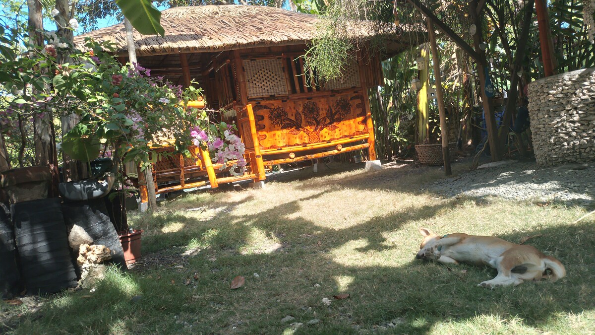 A rustic wooden structure with a thatched roof is set amidst a lush garden. Vibrant flowers bloom near the entrance, while a dog rests peacefully on the sunlit grass. Natural elements and a tranquil atmosphere are present throughout the space.