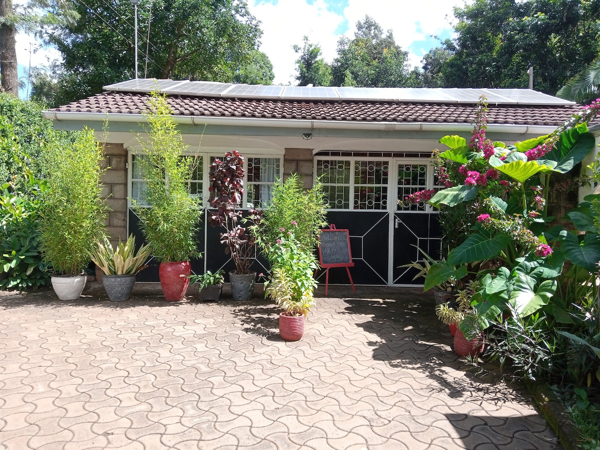 The entrance to the house is framed by lush greenery and vibrant potted plants. A spacious driveway is paved with bricks, leading to the front door. Solar panels are visible on the roof, integrating sustainability with the inviting appearance of the home.