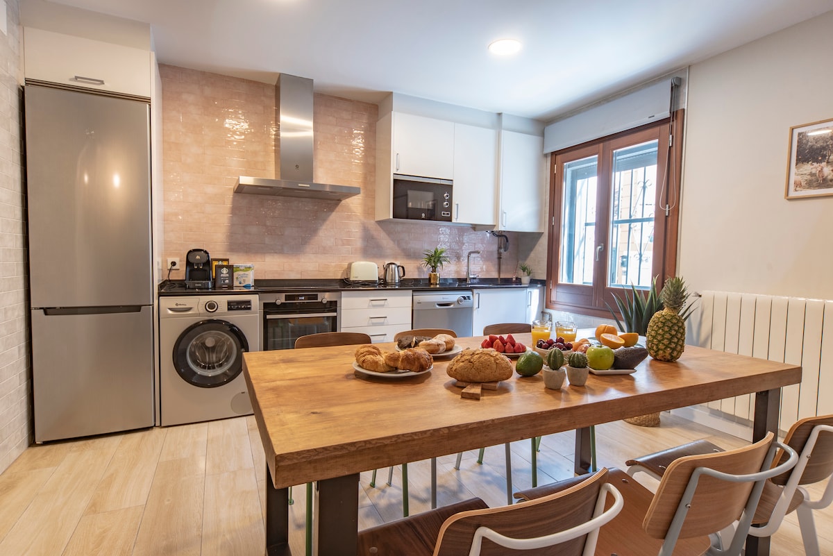 The kitchen and dining area features modern appliances, including a washer and dryer, alongside a spacious wooden table set with a variety of fruits and baked goods. Natural light enters through large windows, illuminating the inviting interior.