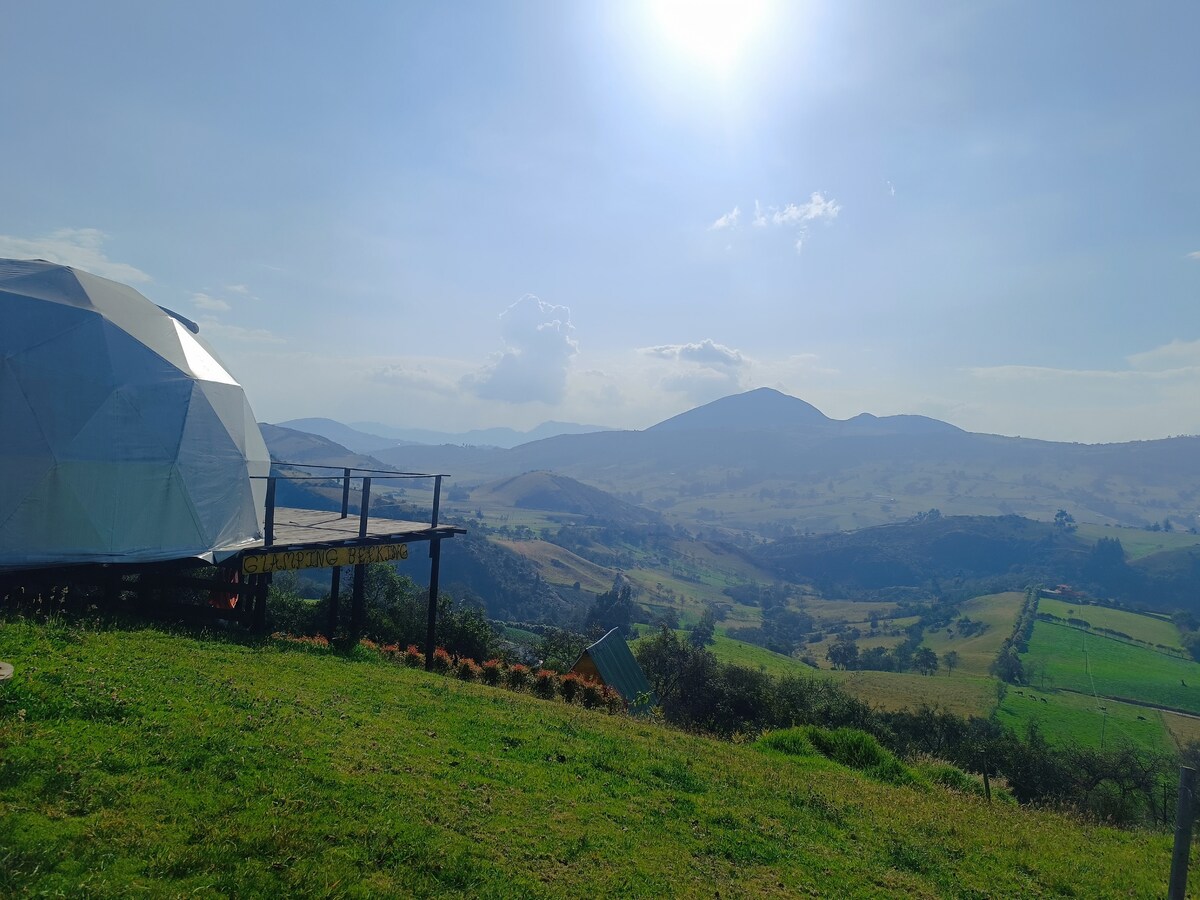 A geodesic dome is positioned on an elevated grassy area, offering expansive views of rolling hills and valleys. The sky is bright with sunlight, casting a gentle glow over the landscape. Lush greenery extends towards the horizon, with distant mountains visible in the background.