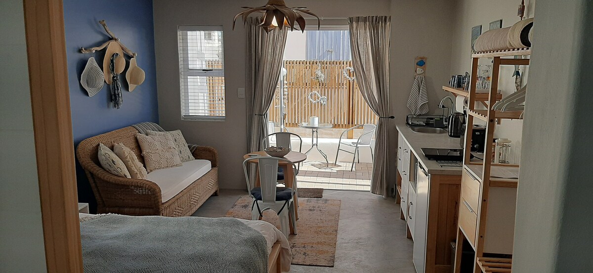 An open-plan living area features a light color scheme with a cozy sofa and a small dining table. Natural light floods in through a large window leading to a private patio. A compact kitchenette is visible, providing essential appliances for convenience.