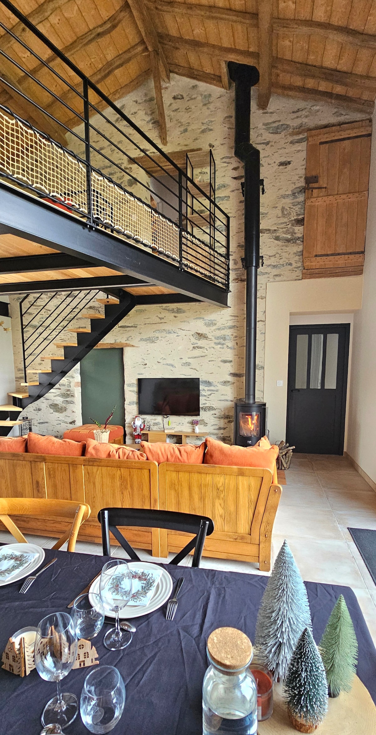 An open living space features a high wooden ceiling with exposed beams and a modern wood stove. A cozy seating area with orange cushions faces a TV, while a dining table set with glassware and decorative trees is visible. A black spiral staircase leads to an upper level.