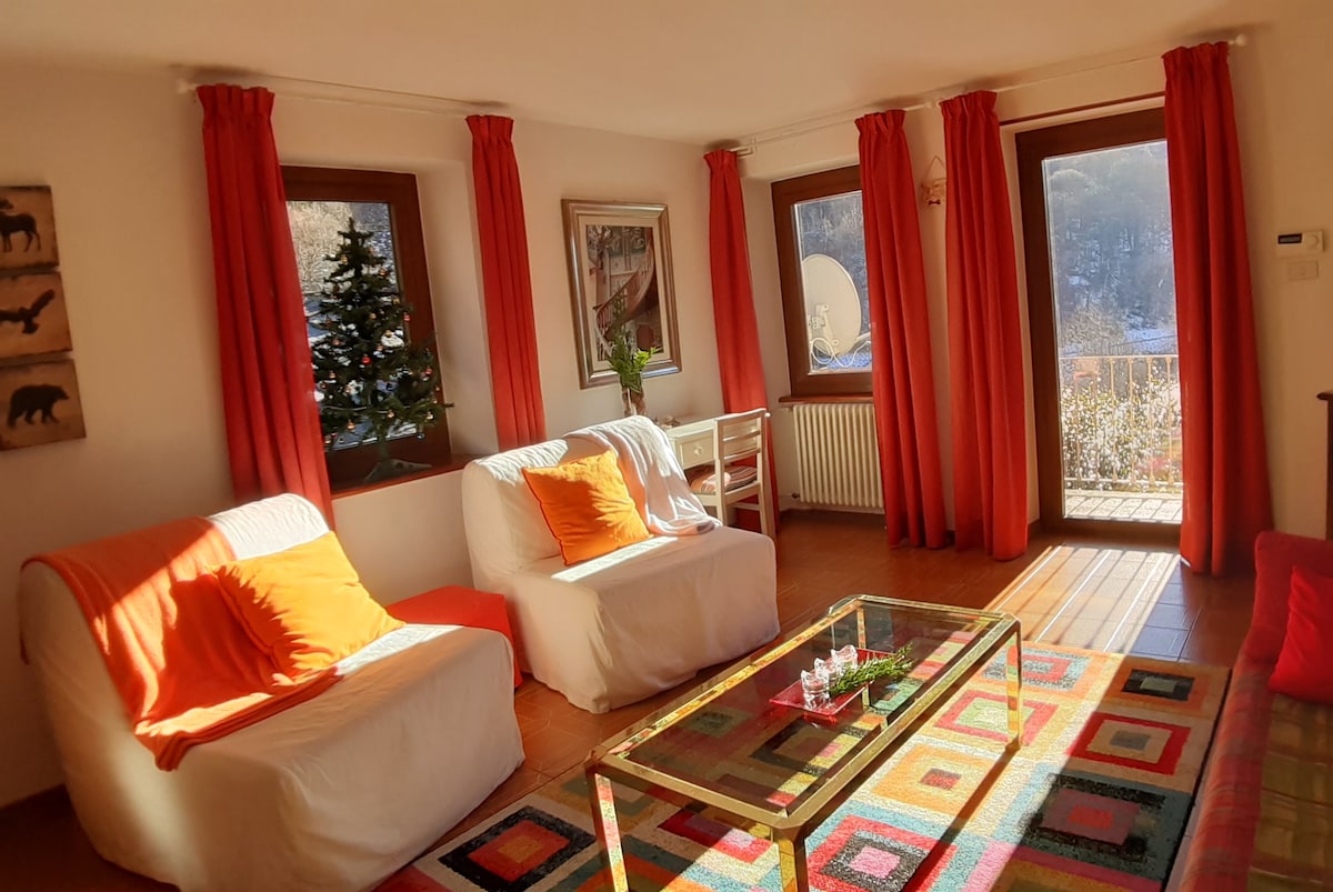 A bright living area features two white upholstered sofas adorned with orange cushions. A glass coffee table sits atop a colorful patterned rug. Large windows with orange curtains allow natural light to illuminate the space, and a small tree is visible in the corner.