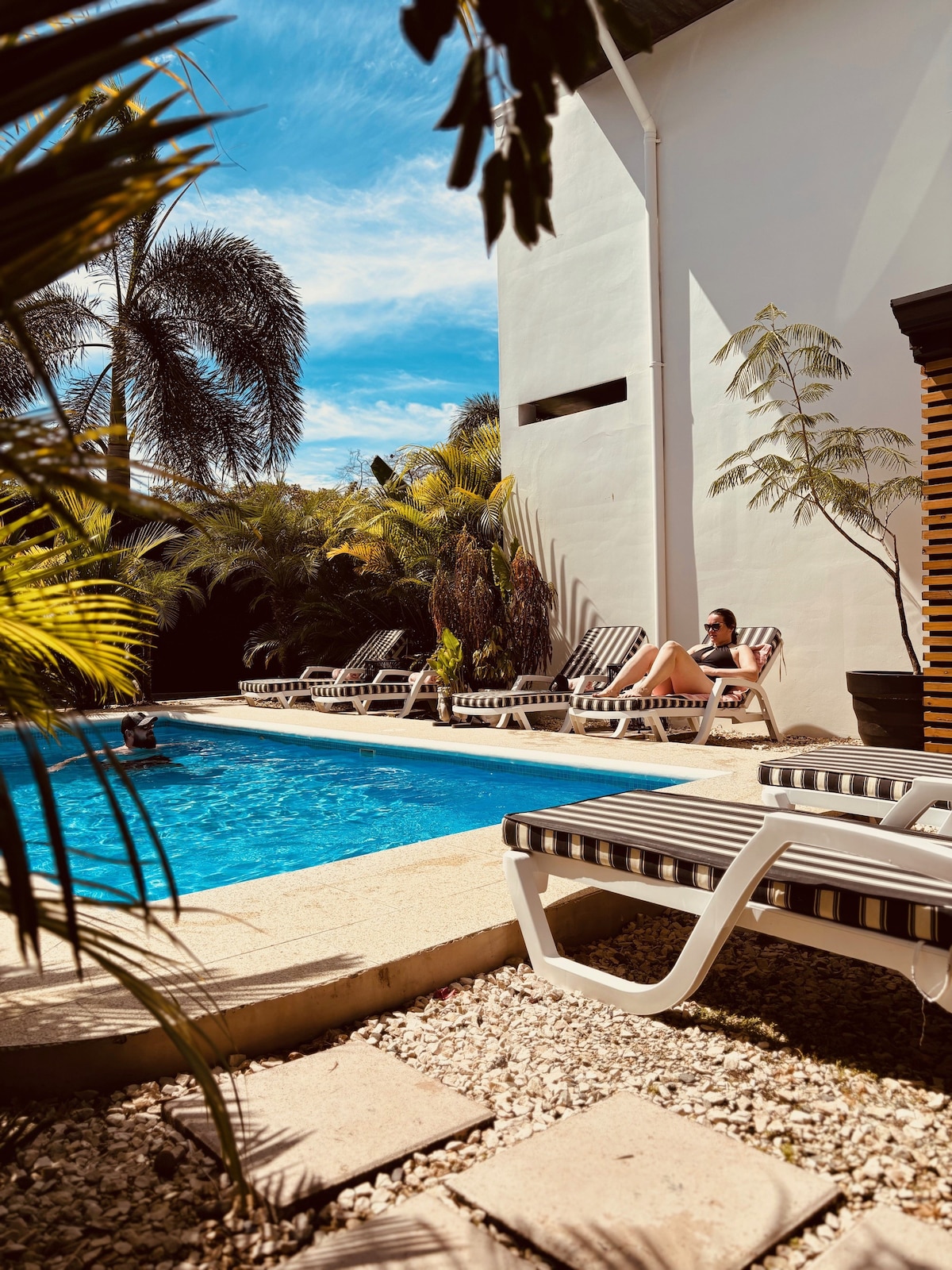 The pool area is surrounded by lush tropical plants, with lounge chairs available for relaxation. A swimmer enjoys the refreshing water in the inviting pool. The sunlit scene showcases a bright blue sky overhead, enhancing the tranquil atmosphere.