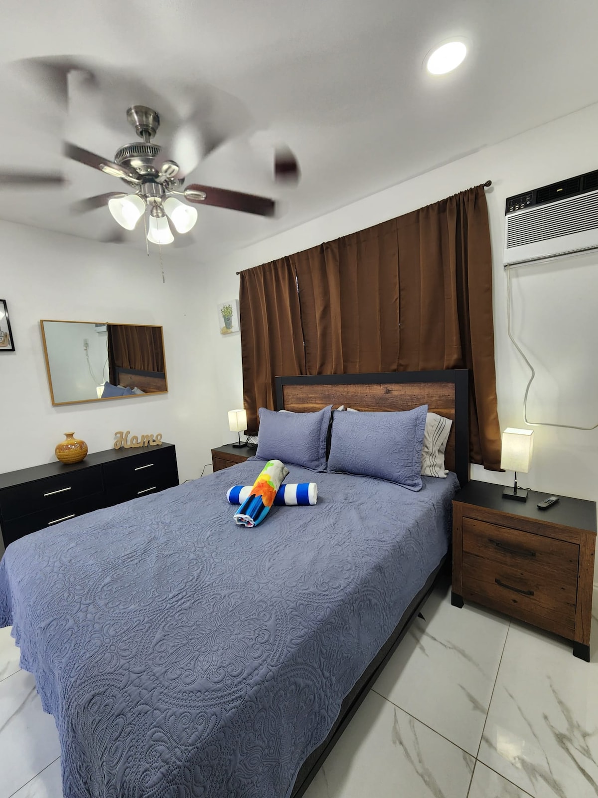 A cozy bedroom features a spacious bed adorned with a blue quilt and decorative towels. Nightstands are positioned on either side, each illuminated by soft lamps. A large mirror reflects the room, and dark curtains frame the window, enhancing the overall decor.