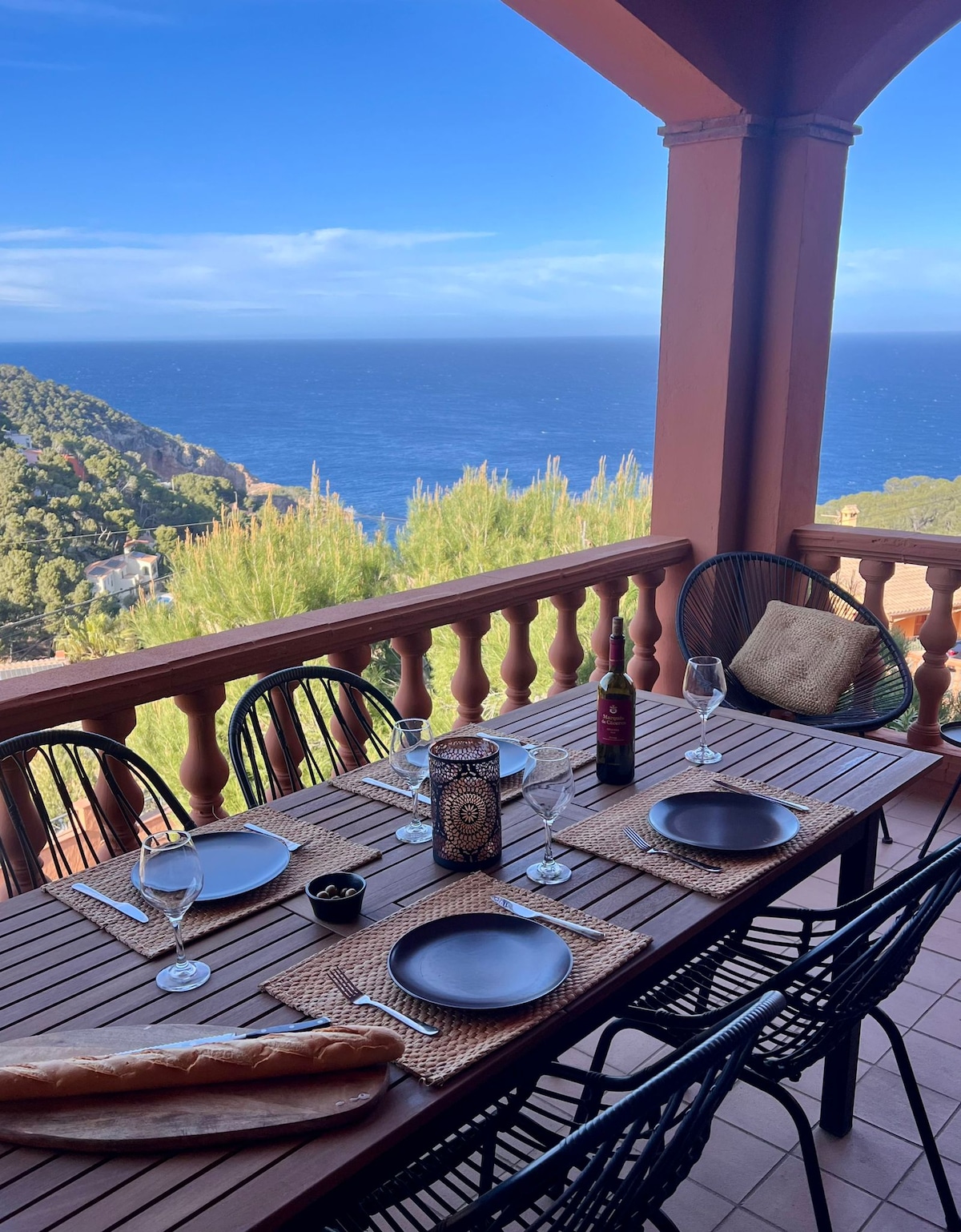An outdoor dining space features a wooden table set for six, with dark woven chairs arranged around it. Plates and glassware are neatly placed along with a decorative centerpiece. Coastal views can be seen in the background, framed by lush greenery.
