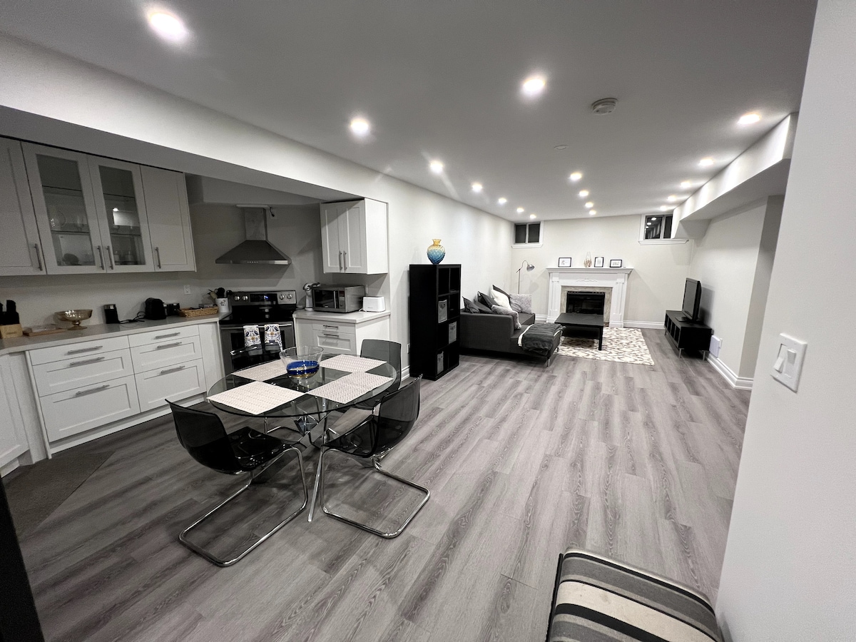 A modern living space is shown, featuring a dining area with a glass-top table and black chairs. A cozy seating area with a sectional sofa is visible, along with a television and indoor fireplace. Bright recessed lighting illuminates the room, enhancing the contemporary design.