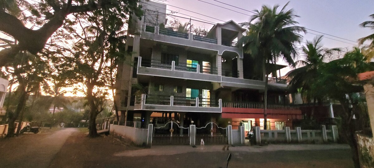 The three-story building is set among lush greenery and palm trees, with balconies visible on each floor. Large windows illuminate the spaces with soft light, while a fence delineates the property from the street, which features ample parking space nearby.