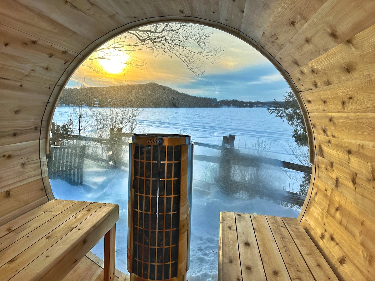 A round wooden sauna interior is visible, with panoramic views through the arch-shaped window. The scene showcases a tranquil winter landscape, featuring snow-covered terrain and a frozen lake. A wooden heater occupies the center, surrounded by spacious wooden benches for seating.