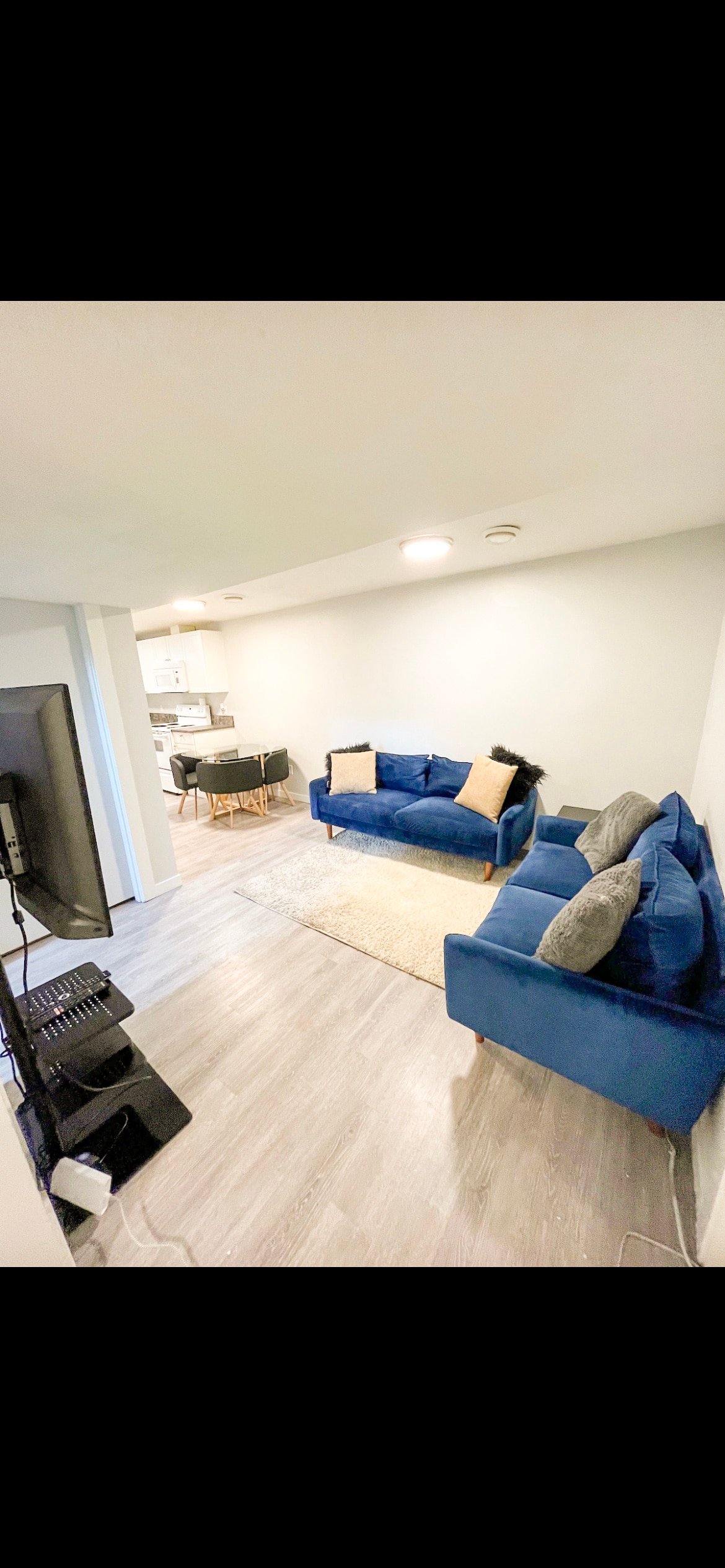A cozy living area features two blue sofas, arranged to create a welcoming space. A light-colored rug lies on the floor, complementing the neutral walls. A television set is positioned nearby, with a dining area visible in the background.