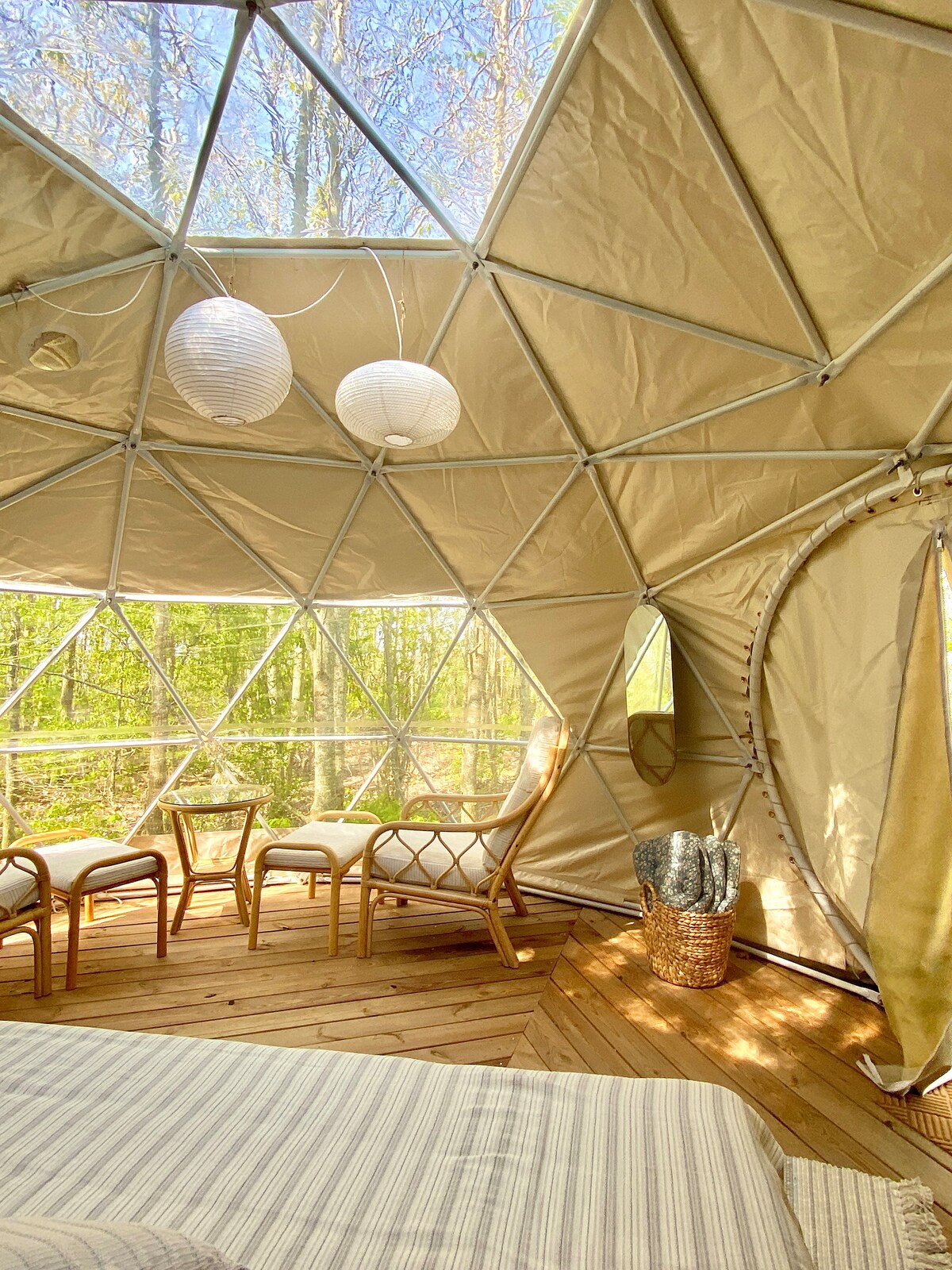 Inside the dome tent, light floods the space through large geometric windows surrounded by greenery. A spacious bed is visible in the foreground, with a cozy seating area featuring two chairs and a small table. Soft lanterns hang from the ceiling, adding warmth.