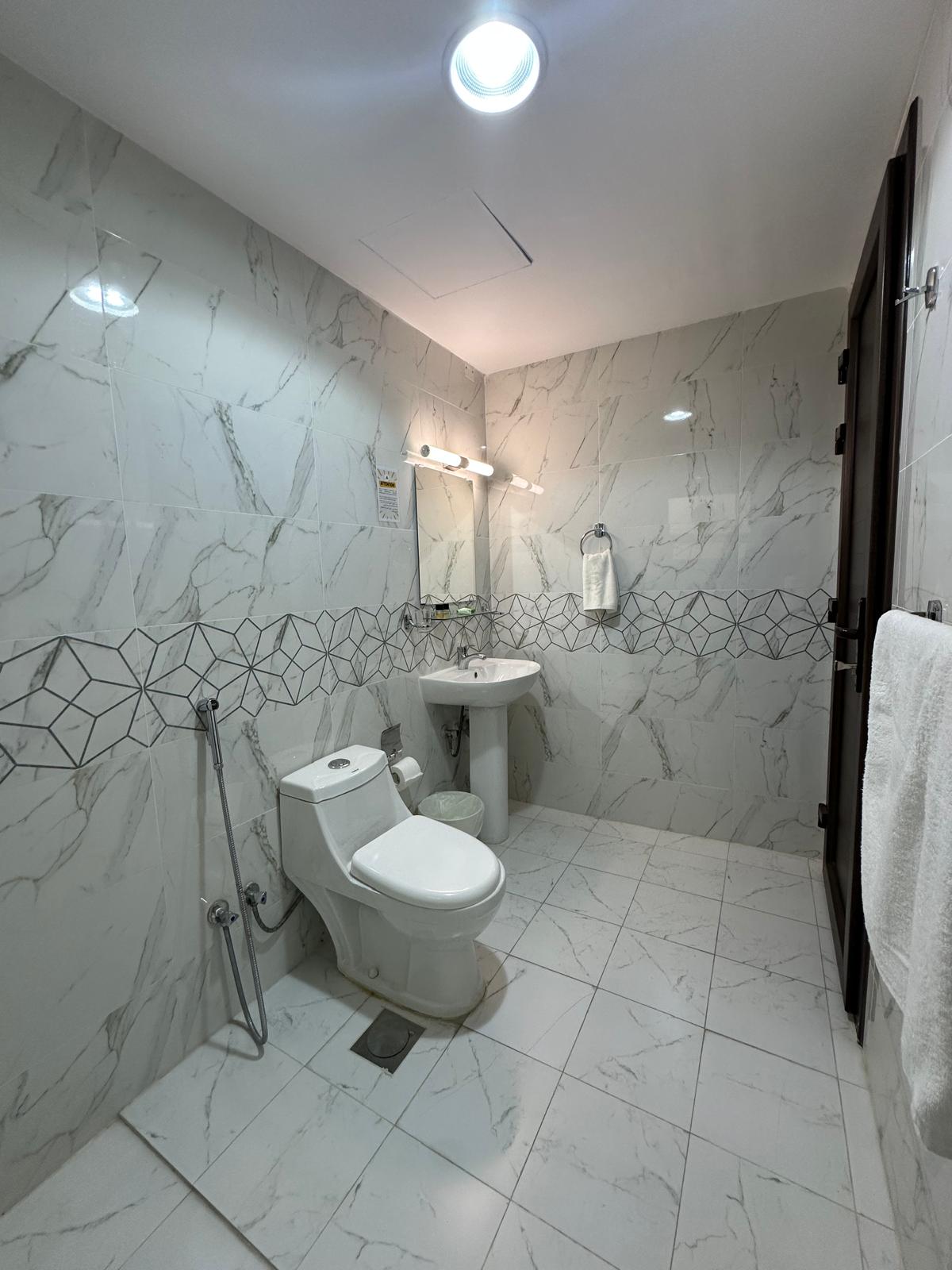 A modern bathroom features sleek white marble walls and tile flooring. A white toilet and a pedestal sink are neatly arranged, with a wall-mounted mirror above the sink. Soft lighting illuminates the space, enhancing the clean and fresh atmosphere.