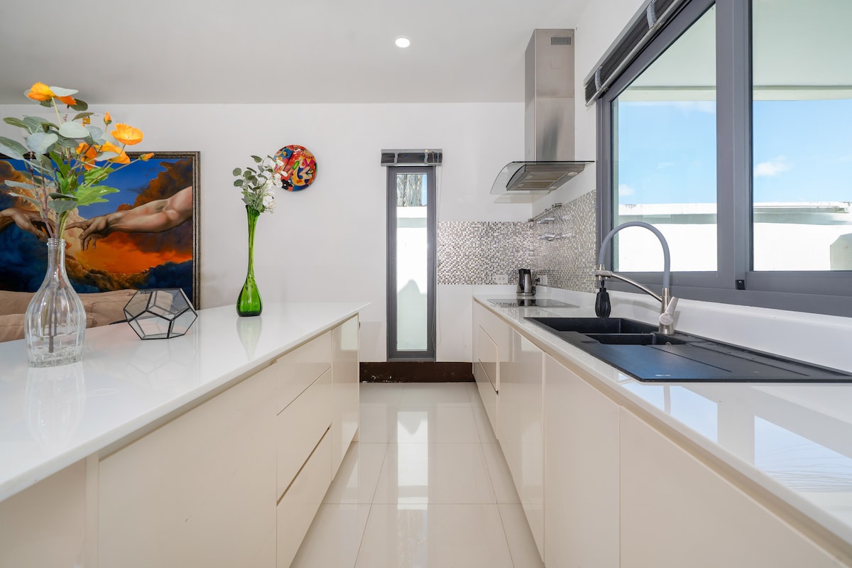 The modern kitchen features sleek white cabinetry and a spacious countertop. A stainless steel sink and stove are present, accompanied by a stylish backsplash. Natural light enters through large windows, illuminating decorative elements including a vase with flowers and a colorful wall art piece.