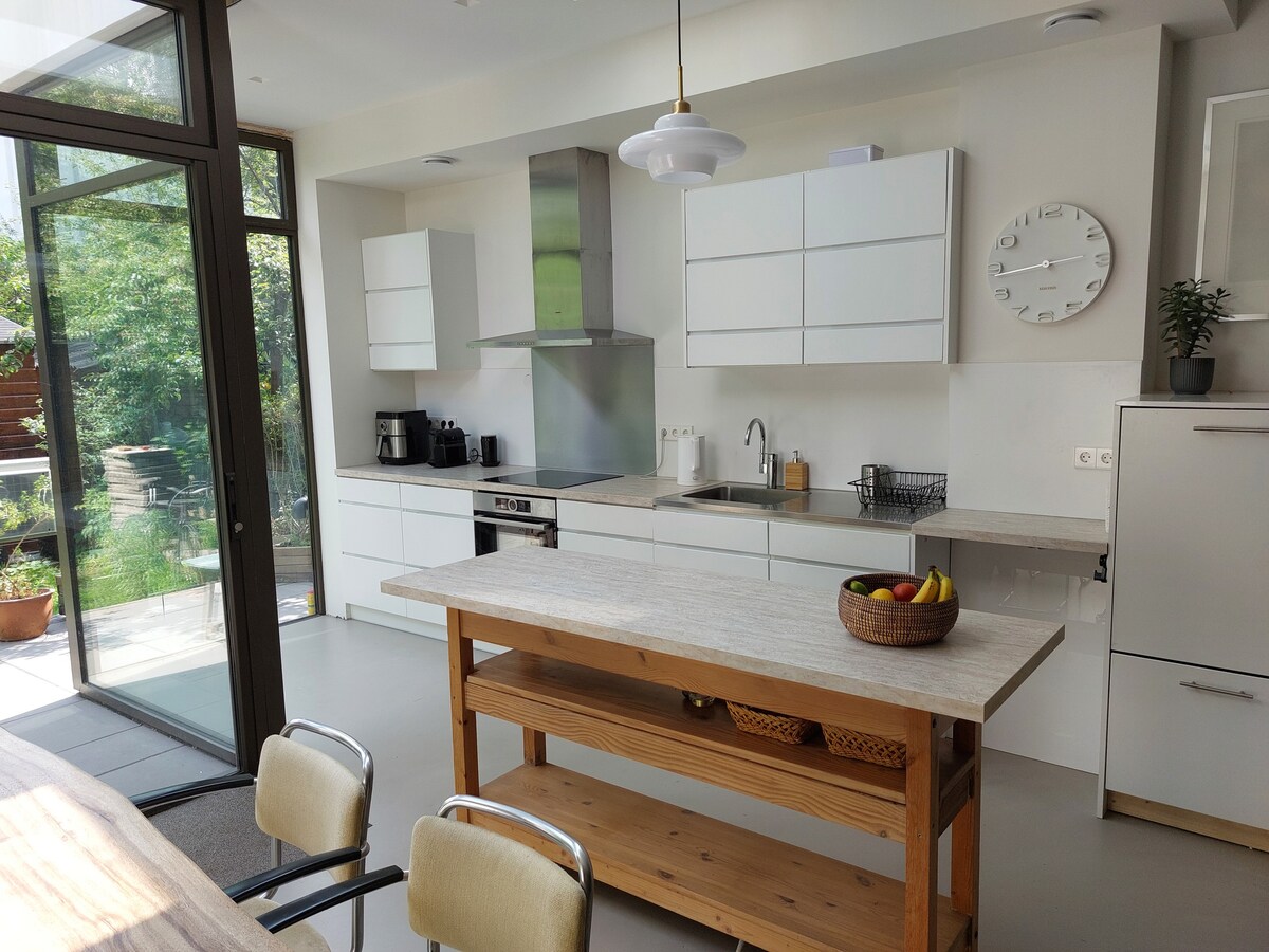 The kitchen features modern cabinetry with a minimalist design, complemented by a central wooden island. A stainless-steel range hood and appliances reflect natural light from large glass doors leading to the garden. A bowl of fresh fruits adds a touch of color to the countertop.