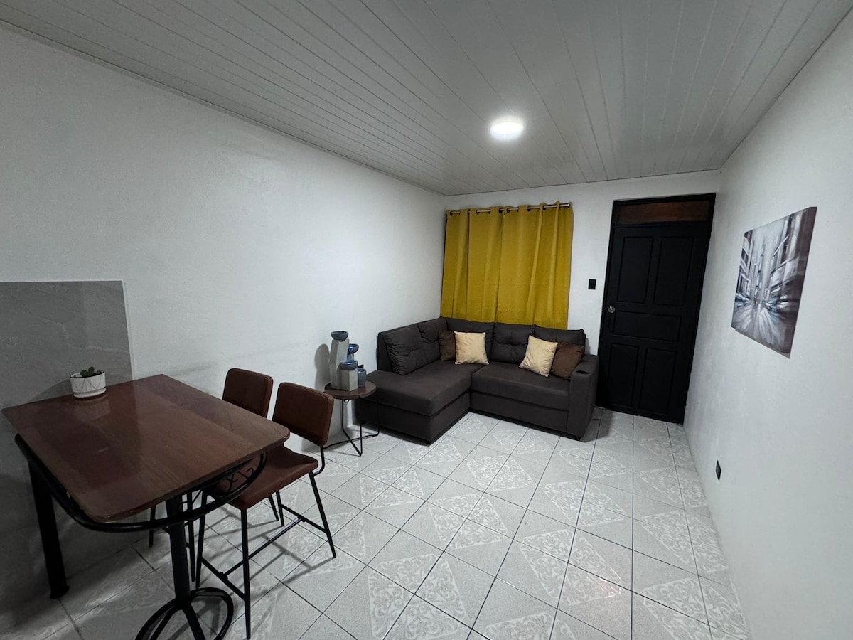 A cozy living area features a brown sectional sofa with yellow accent pillows. A wooden dining table with four chairs is positioned nearby. Light-colored tile flooring complements bright white walls, and a window with yellow curtains allows natural light to fill the space.