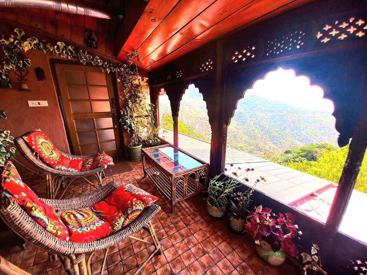 A serene balcony is presented with ornate wooden arches, offering views of the surrounding mountains. Two comfortable wicker chairs with vibrant cushions are positioned beside a glass-topped table, accompanied by potted plants that enhance the natural ambiance.