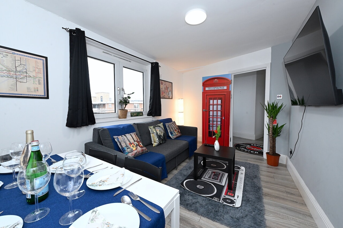 The living room features a modern sofa with decorative pillows, a sleek coffee table, and a playful red telephone booth. Two large windows allow natural light to fill the space, while a dark rug enhances the room's inviting atmosphere. A television is mounted on the wall opposite the seating area.