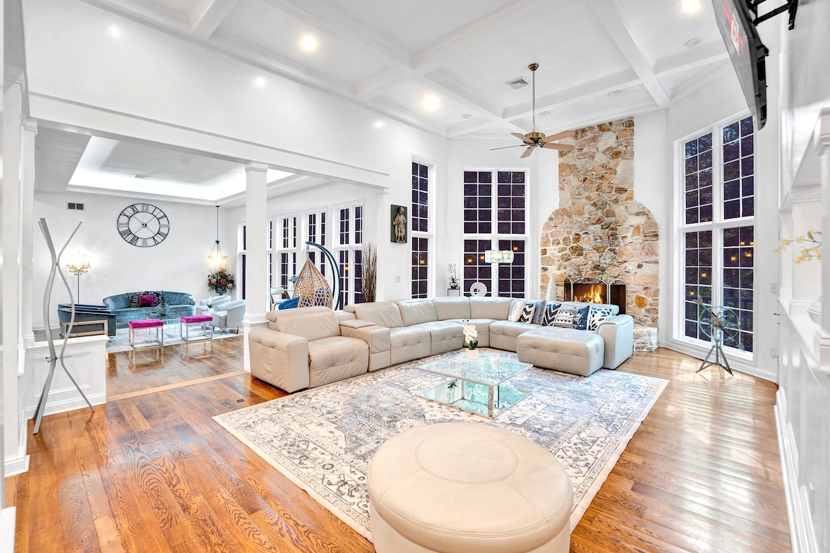 A spacious living area features a large seating arrangement with plush sofas, surrounding a glass coffee table atop a decorative area rug. Natural light fills the space through tall windows, highlighting a stone fireplace and elegant decor throughout.
