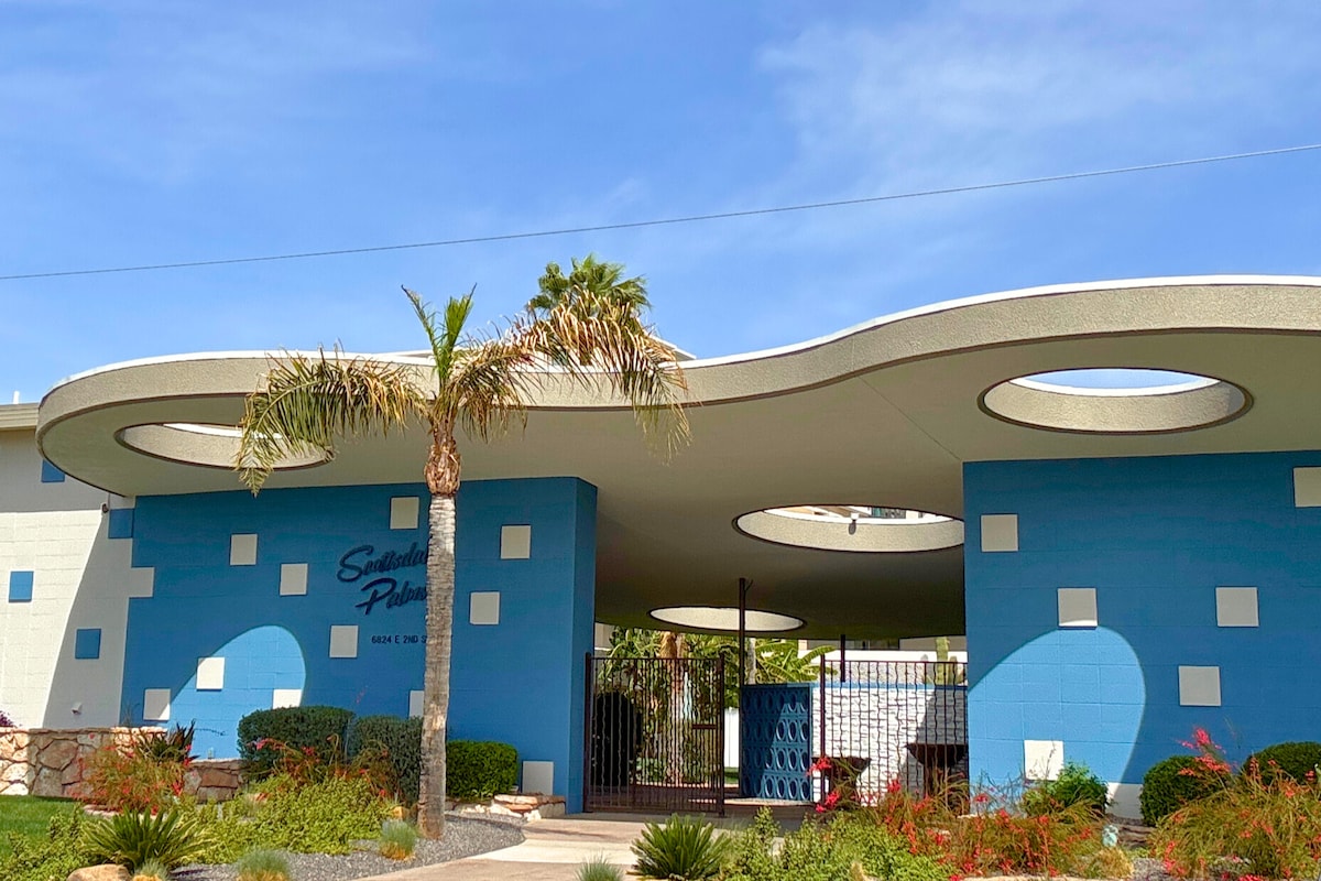A mid-century condominium building is showcased, featuring googie-style rooflines with circular cutouts. The walls are painted in vibrant blue, complemented by geometric white accents. Lush gardens and palm trees enhance the outdoor space, creating a welcoming entrance to the community.