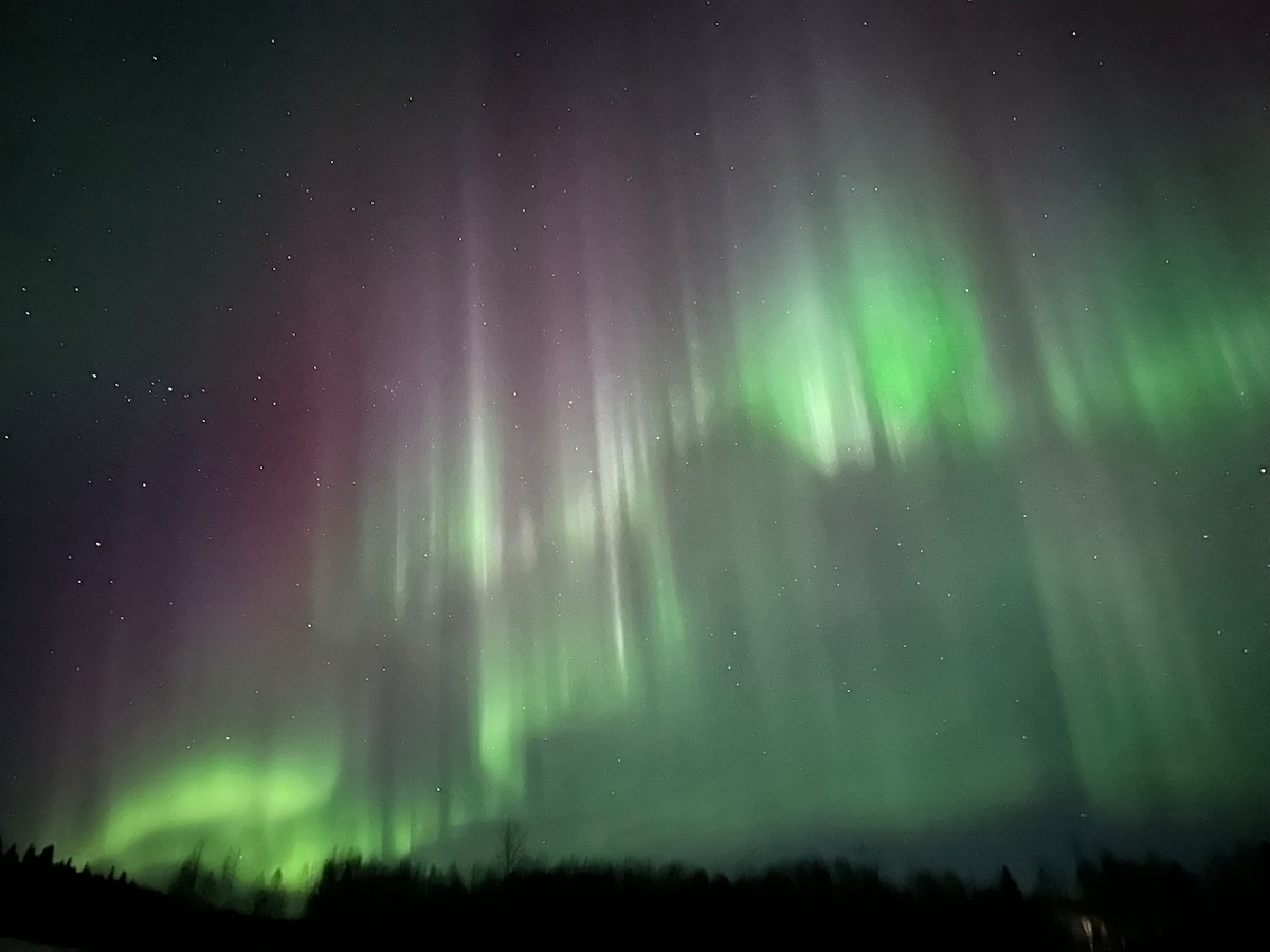 The night sky is illuminated by vibrant auroras, with shades of green, purple, and hints of blue creating a dynamic display. Stars are faintly visible, contributing to the serene atmosphere of the wilderness landscape below.