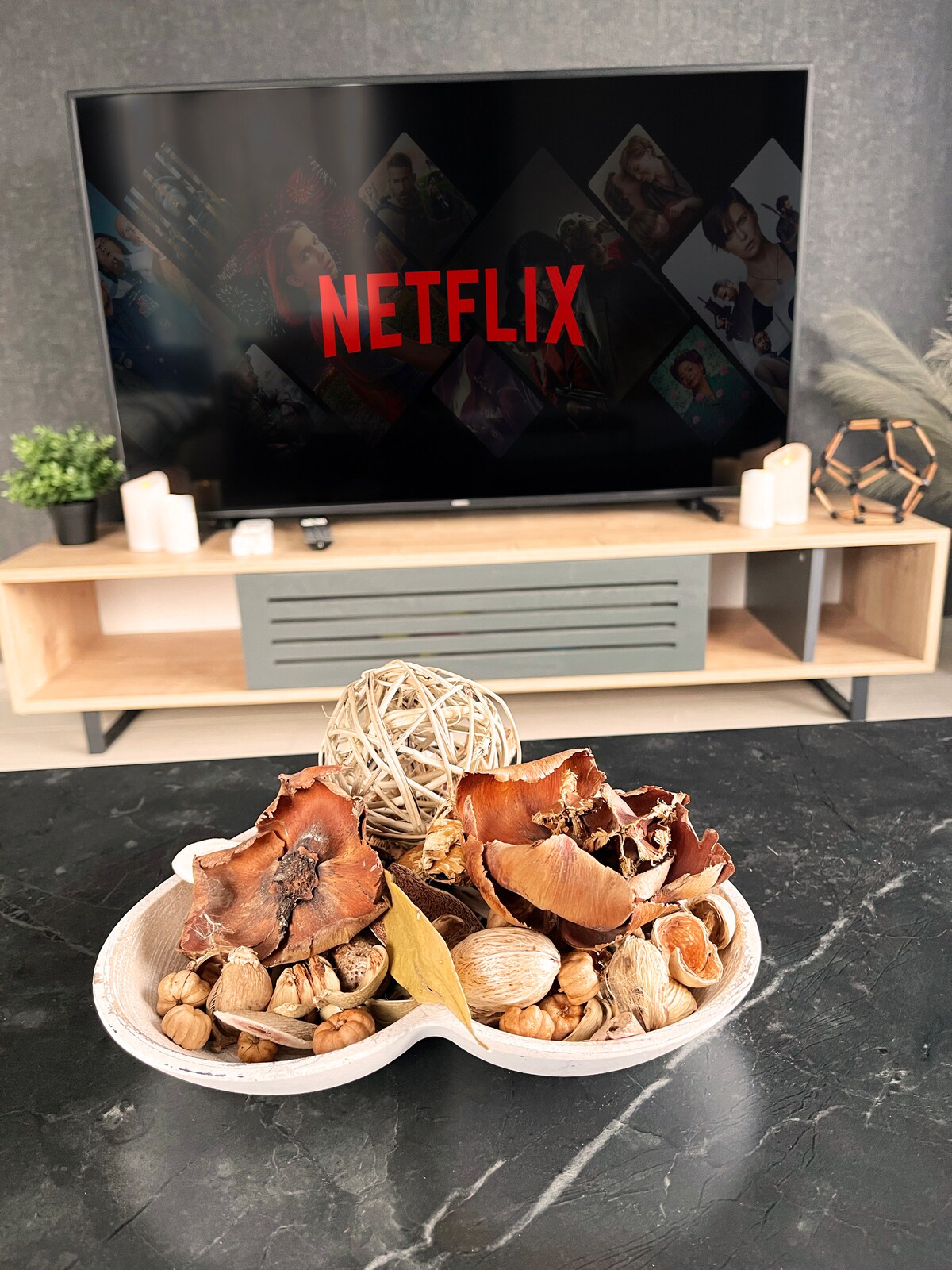 A decorative bowl filled with natural elements such as shells and dried leaves is positioned in front of a sleek entertainment unit. A large television displaying the Netflix logo serves as a focal point, enhancing the inviting design of the living space.