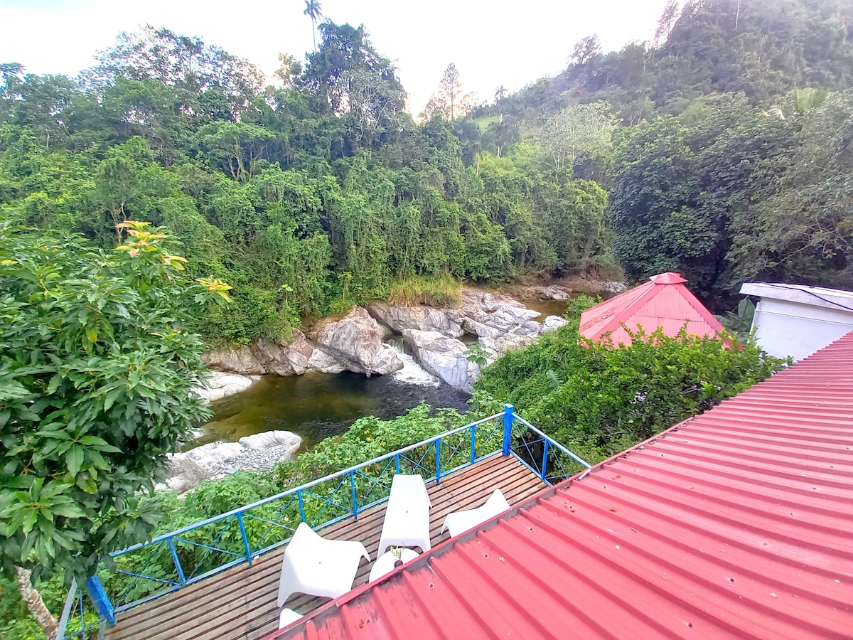 An outdoor terrace with white chairs provides a scenic view of the Mahomita River, flanked by rocky banks and lush greenery. The surrounding mountains are visible in the background, enhancing the natural setting. A red roof structure is partially visible in the image.