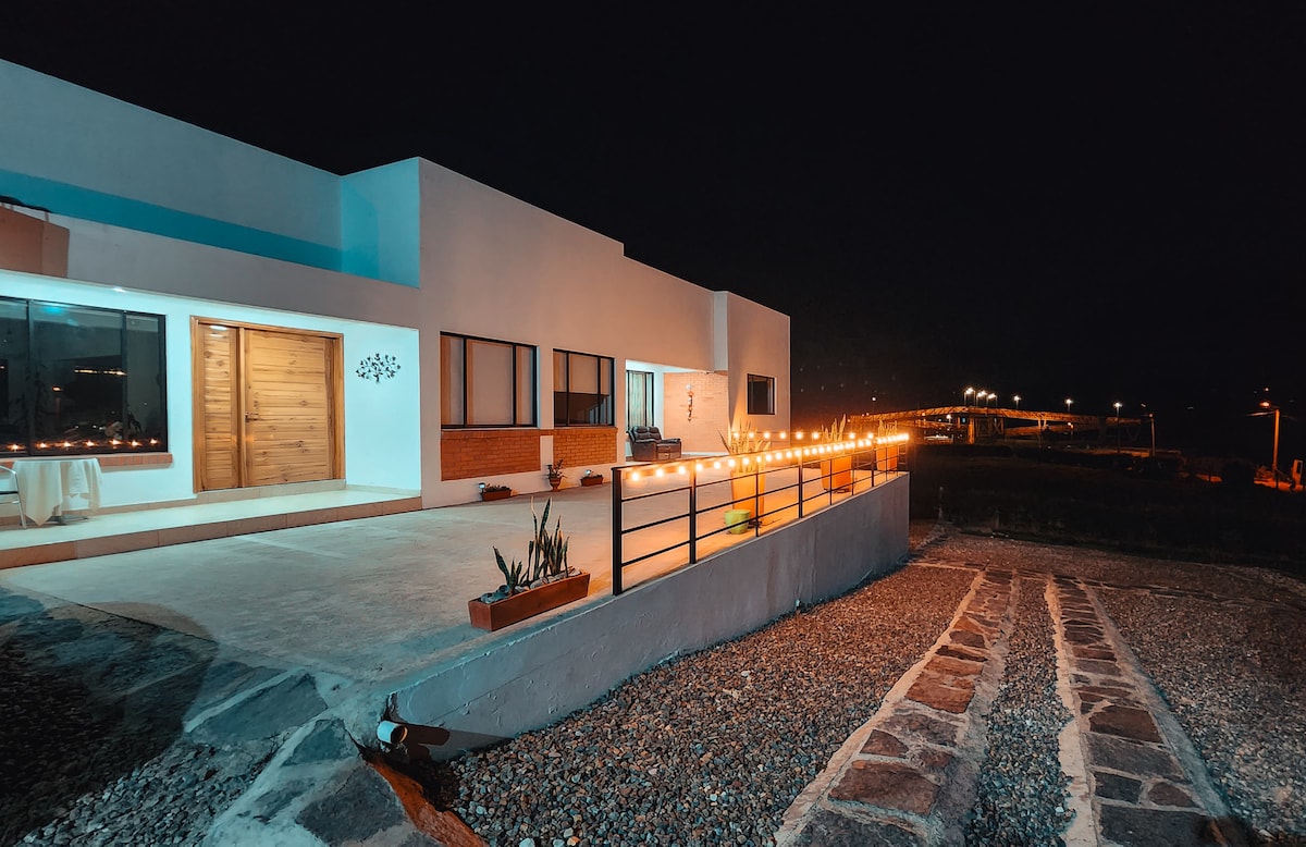A modern structure is shown at night, with outdoor lighting creating a warm glow. A spacious patio is visible, accompanied by a smooth stone pathway leading to the entrance. Large windows allow for a view of the surroundings, while the night sky provides a serene backdrop.